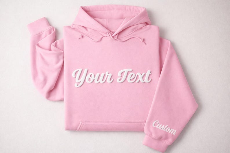 May include: A light pink hoodie with a drawstring hood and a kangaroo pocket. The words "Your Text" are displayed in a white, cursive font across the chest. The right sleeve has the word "Custom" in white.
