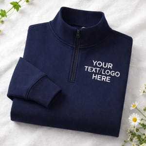 Puede incluir: Sudadera azul marino con cremallera de un cuarto y el texto "YOUR TEXT/LOGO HERE" en blanco. La sudadera tiene una cremallera negra y puños acanalados. La prenda se muestra sobre una superficie blanca con pequeñas flores blancas.