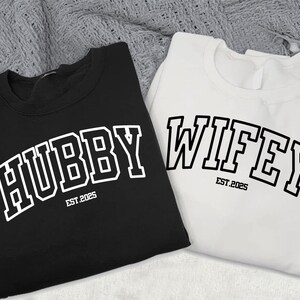 May include: Two black and white sweatshirts with the words "Hubby Est. 2025" and "Wifey Est. 2025" printed on them in a collegiate font.