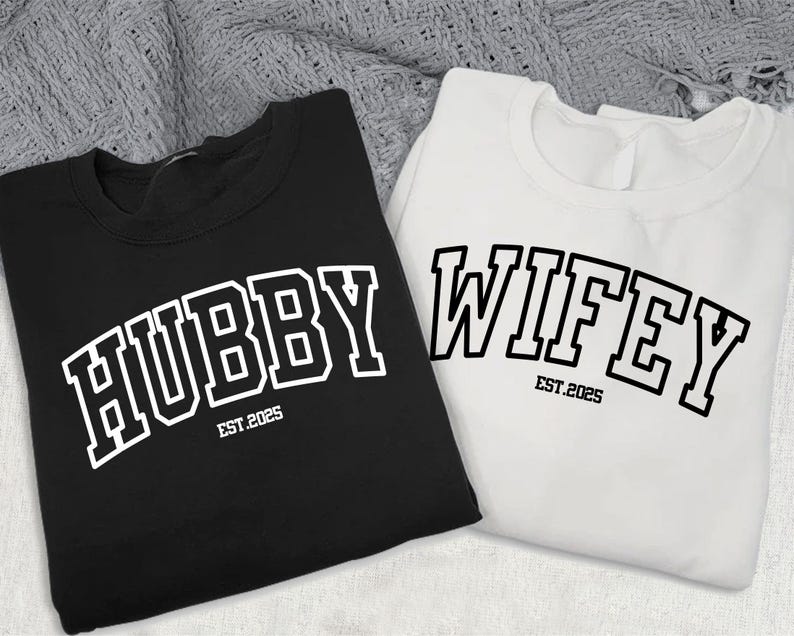 May include: Two black and white sweatshirts with the words "Hubby Est. 2025" and "Wifey Est. 2025" printed on them in a collegiate font.
