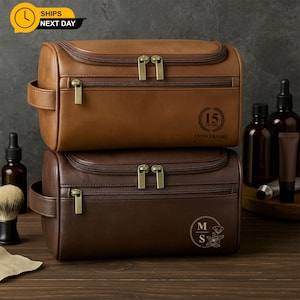 May include: Two leather wash bags, one in brown and one in dark brown, stacked. The top bag has "15 YEARS ANNIVERSARY" printed on it. Both have gold zippers and handles. Other grooming products are visible.
