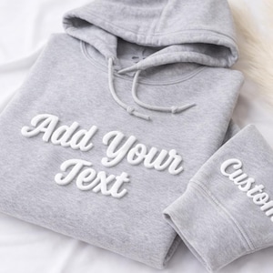 May include: A light gray hoodie and matching cuff with the words "Add Your Text" and "Custom" in white, cursive lettering. The hoodie has a drawstring and hood. The items are on a white surface.