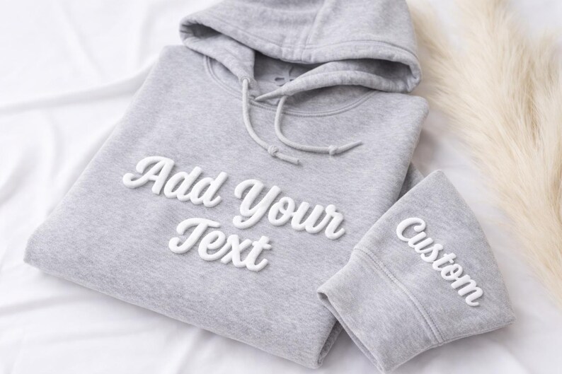 May include: A light gray hoodie and matching cuff with the words "Add Your Text" and "Custom" in white, cursive lettering. The hoodie has a drawstring and hood. The items are on a white surface.