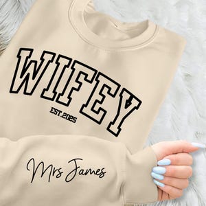 May include: A beige sweatshirt with the text "WIFEY EST. 2025" in black outlined letters. The text "Mrs James" is written in cursive below the larger text.