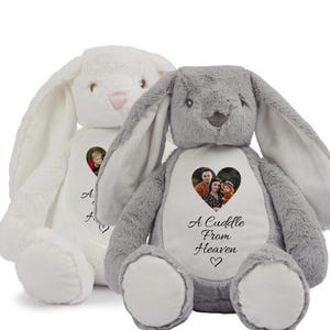 May include: Two plush bunny rabbits, one white and one grey, with a heart-shaped photo frame on their chests. The grey bunny has the text "A Cuddle From Heaven" printed below the photo frame.