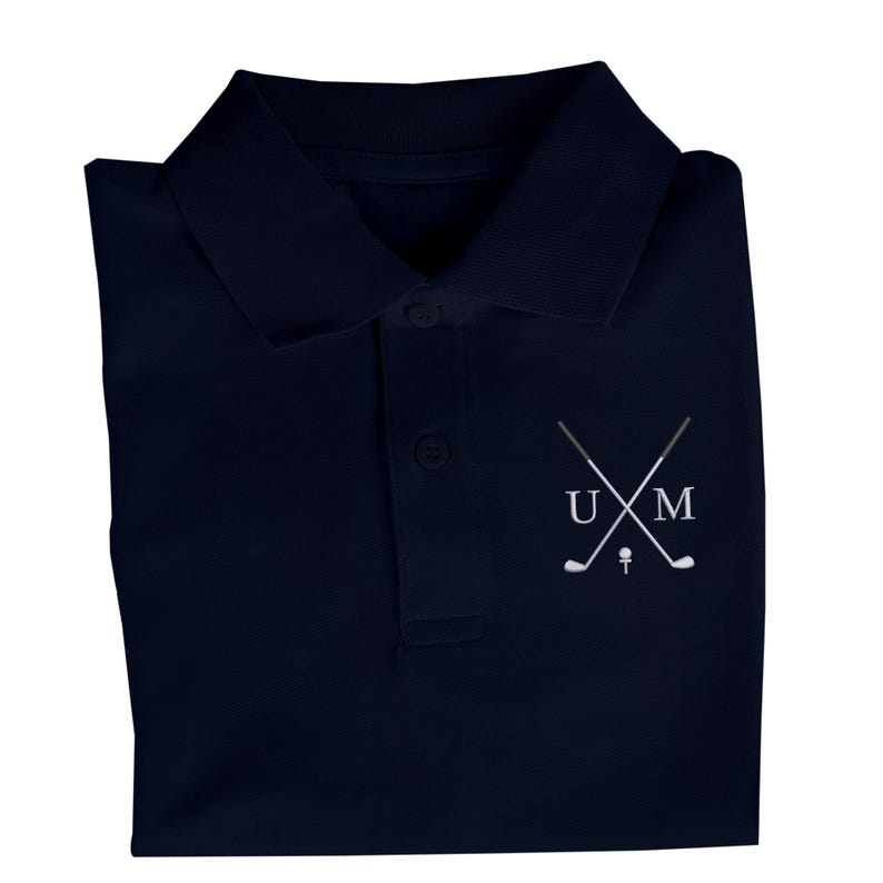 K&ouml;nnte beinhalten: Ein marineblaues Poloshirt mit einem wei&szlig;en gestickten Logo, das gekreuzte Golfschl&auml;ger und die Buchstaben "UM" zeigt.