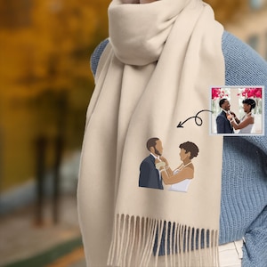 May include: A cream-coloured scarf with a fringed edge, featuring an embroidered illustration of a couple. A small photo of the couple is also included. The scarf is draped over a person wearing a blue jumper. The background is blurred.