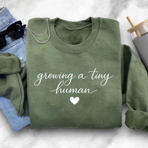 May include: An olive green sweatshirt with the text "growing a tiny human" in white script and a white heart. The sweatshirt is paired with blue jeans, black sunglasses, a silver necklace, and a grey tumbler with a wooden straw.