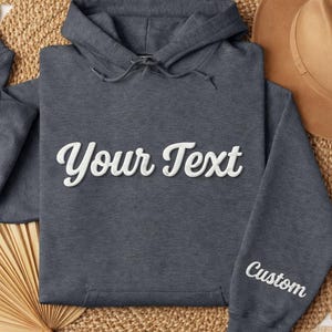 May include: A dark gray hoodie with the text "Your Text" in white script across the chest. The word "Custom" is embroidered on the sleeve. The hoodie is displayed on a woven surface with a brown hat.