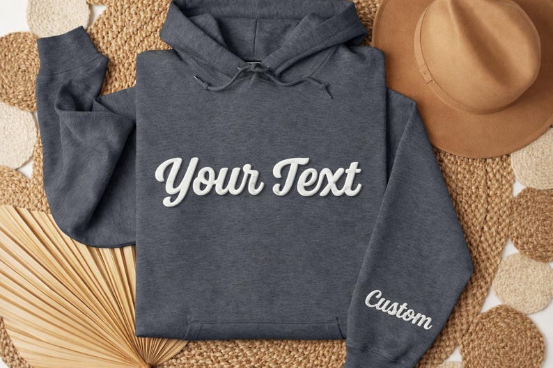 May include: A dark gray hoodie with the text "Your Text" in white script across the chest. The word "Custom" is embroidered on the sleeve. The hoodie is displayed on a woven surface with a brown hat.