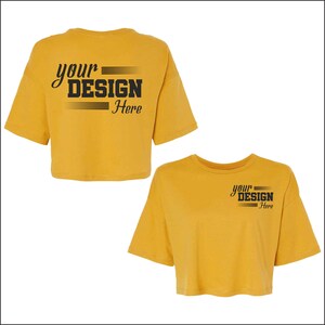 May include: Mustard yellow crop top t-shirts. One shirt has the text "your DESIGN Here" on the back. The other shirt has the same text on the front. Both shirts have short sleeves and a crew neck.