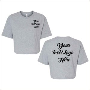 May include: Two light gray crop tops. The front of the shirt has the text "Your text/Logo Here" in black script. The back of the shirt has the same text. The shirts have short sleeves and a crew neck.