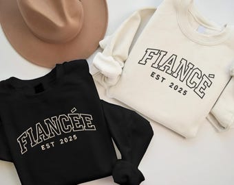 May include: Two black and white sweatshirts with the text "Fiancé Est 2025" and "Fiancée Est 2025" in a varsity style font.