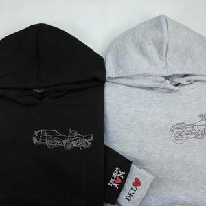 Puede incluir: Dos sudaderas con capucha, una negra y otra gris, con dise&ntilde;os bordados. La sudadera negra tiene un dise&ntilde;o de coche y moto, mientras que la sudadera gris tiene un dise&ntilde;o de moto. La sudadera gris tambi&eacute;n tiene un peque&ntilde;o coraz&oacute;n rojo con el texto "DKL A&hearts;M 3.25.2022" en la manga.