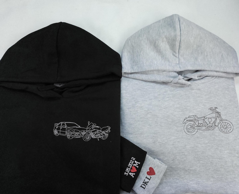 Puede incluir: Dos sudaderas con capucha, una negra y otra gris, con dise&ntilde;os bordados. La sudadera negra tiene un dise&ntilde;o de coche y moto, mientras que la sudadera gris tiene un dise&ntilde;o de moto. La sudadera gris tambi&eacute;n tiene un peque&ntilde;o coraz&oacute;n rojo con el texto "DKL A&hearts;M 3.25.2022" en la manga.