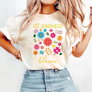 May include: A white t-shirt with a floral design and the text "Let Kindness Bloom" in yellow lettering.