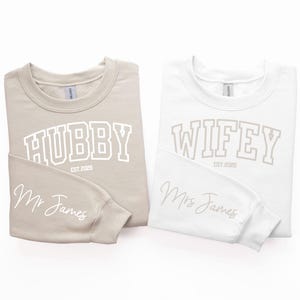 May include: Two long-sleeved sweatshirts, one beige and one white, with the words "Hubby Est 2023" and "Wifey Est 2023" printed on the front. The beige sweatshirt has "Mr. James" written on the sleeve, and the white sweatshirt has "Mrs. James" written on the sleeve.