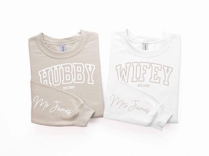 May include: Two long-sleeved sweatshirts, one beige and one white, with the words "Hubby Est 2023" and "Wifey Est 2023" printed on the front. The beige sweatshirt has "Mr. James" written on the sleeve, and the white sweatshirt has "Mrs. James" written on the sleeve.