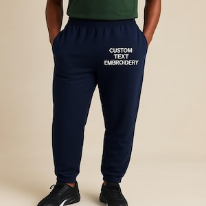 Puede incluir: Pantalones deportivos azul marino oscuro con el texto "CUSTOM TEXT EMBROIDERY" en blanco. Los pantalones tienen bolsillos y cintura con cordón. La persona de la imagen lleva zapatillas negras.