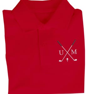 K&ouml;nnte beinhalten: Rotes Poloshirt mit einem wei&szlig;en gestickten Design aus zwei gekreuzten Golfschl&auml;gern und den Buchstaben "UM" dar&uuml;ber.
