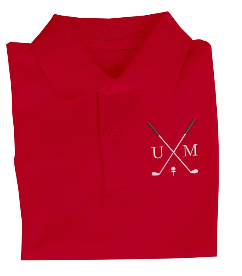 K&ouml;nnte beinhalten: Rotes Poloshirt mit einem wei&szlig;en gestickten Design aus zwei gekreuzten Golfschl&auml;gern und den Buchstaben "UM" dar&uuml;ber.