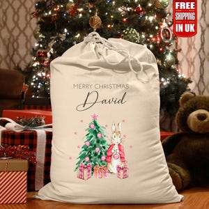 Può includere: Un sacco di Babbo Natale in tela bianca con il nome personalizzato "David" e un albero di Natale con un coniglio che tiene un regalo. Il sacco ha la scritta "Buon Natale" stampata.