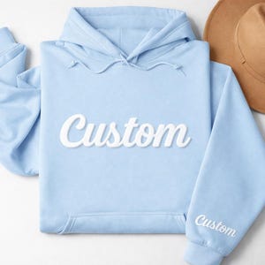 May include: A light blue hoodie with the word "Custom" in white script across the front. The sleeve also has the word "Custom" embroidered in white. A tan fedora hat is in the background.