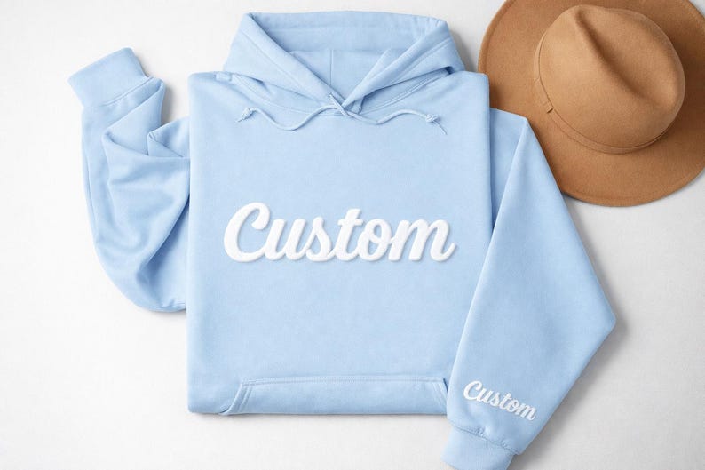 May include: A light blue hoodie with the word "Custom" in white script across the front. The sleeve also has the word "Custom" embroidered in white. A tan fedora hat is in the background.