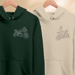 Puede incluir: Dos sudaderas con capucha, una verde y otra beige, con un dibujo lineal de una motocicleta en cada una. La sudadera verde tiene una motocicleta sin conductor, mientras que la sudadera beige tiene una motocicleta con un conductor.