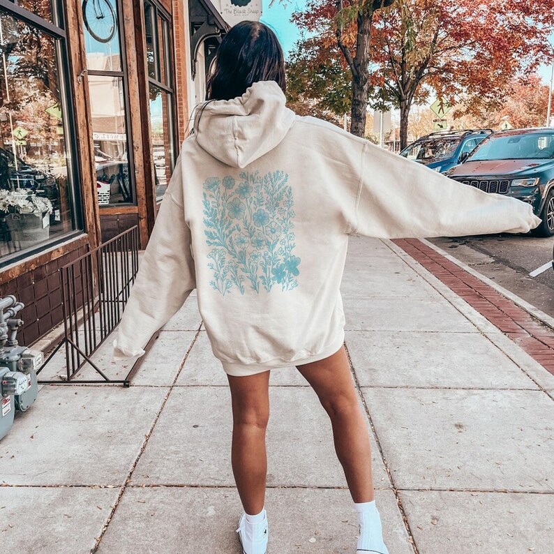 May include: A cream-colored hoodie with a blue floral print on the back. The hoodie has a drawstring hood and a relaxed fit.
