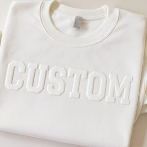 Op de afbeelding: Witte trui met het woord "CUSTOM" in reliëf.