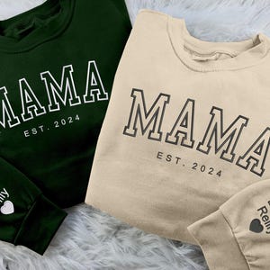 May include: Two sweatshirts are shown, one dark green and one beige. Each has the word "MAMA" in a bold font, with "EST. 2024" underneath. The green sweatshirt's sleeve displays "Daniel Reilly" and a heart. The beige one has the same text.
