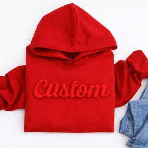 May include: A red hoodie with the word "Custom" embroidered on the front. The hoodie has a hood and long sleeves. A pair of blue jeans is next to the hoodie on a white surface. Apparel.