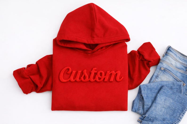 May include: A red hoodie with the word "Custom" embroidered on the front. The hoodie has a hood and long sleeves. A pair of blue jeans is next to the hoodie on a white surface. Apparel.