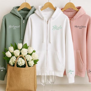 May include: Three zip-up hoodies in sage green, white, and pink, hanging on wooden hangers. The hoodies are embroidered with the words "Bridesmaid", "Bride", and "Maid of Honor", along with dates. A burlap tote bag with white flowers and two wine glasses are in the foreground.