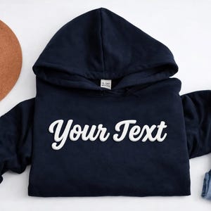 May include: Navy blue hooded sweatshirt with the text "Your Text" in a white, cursive font. A brown hat and a pair of blue jeans are also in the image. The sweatshirt has a drawstring hood and long sleeves.