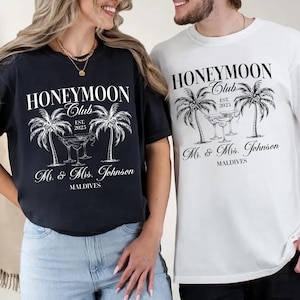 Peut inclure: Un couple portant des t-shirts assortis noir et blanc avec le texte "Honeymoon Club Est. 2025" et des palmiers. Les t-shirts comprennent également le texte "Mr. & Mrs. Johnson Maldives".