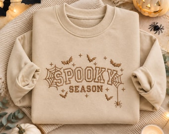 Op de afbeelding: Een beige sweater met ronde hals en bruin borduurwerk met de tekst "SPOOKY SEASON" in een college-lettertype. Het ontwerp is versierd met geborduurde spinnenwebben, kleine vleermuizen, sterretjes en een hangende spin als detail.