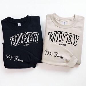 May include: Two sweatshirts, one black and one beige, with the words "Hubby" and "Wifey" in a collegiate font. The text "Est. 2025" appears below each word. The black sweatshirt has the text "Mr. Thomas" on the sleeve, and the beige sweatshirt has the text "Mrs. Thomas" on the sleeve.