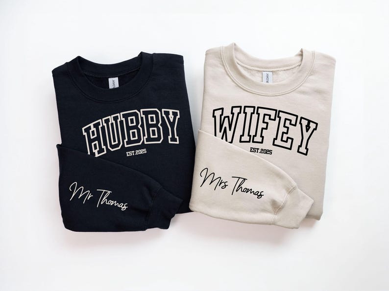 May include: Two sweatshirts, one black and one beige, with the words "Hubby" and "Wifey" in a collegiate font. The text "Est. 2025" appears below each word. The black sweatshirt has the text "Mr. Thomas" on the sleeve, and the beige sweatshirt has the text "Mrs. Thomas" on the sleeve.