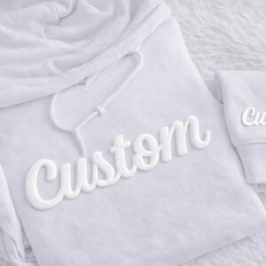 May include: A white hoodie with the word "Custom" in a 3D style across the front. The word "Custom" is also on the sleeve. The hoodie has a drawstring and a hood.