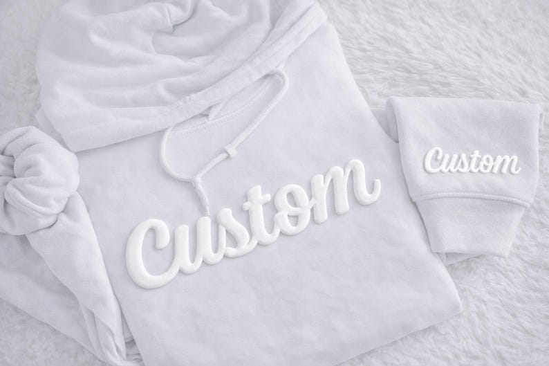 May include: A white hoodie with the word "Custom" in a 3D style across the front. The word "Custom" is also on the sleeve. The hoodie has a drawstring and a hood.