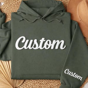 May include: An olive green hoodie with the word "Custom" in white script across the chest and on the sleeve. A brown hat rests nearby on a woven surface. The hoodie has a drawstring hood and a front pocket.