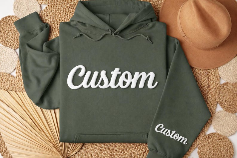 May include: An olive green hoodie with the word "Custom" in white script across the chest and on the sleeve. A brown hat rests nearby on a woven surface. The hoodie has a drawstring hood and a front pocket.