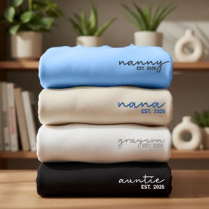 May include: Four folded sweatshirts in a stack, in light blue, beige, white, and black. Each sweatshirt has a different name and the year "EST. 2026" printed on it. The top sweatshirt says "nanny", the second says "nana", the third says "grayson", and the bottom says "auntie".