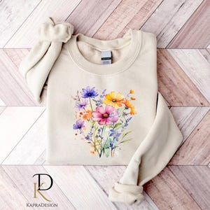 May include: A beige sweatshirt with a watercolor floral design featuring pink, yellow, and purple flowers. The flowers are arranged in a bouquet shape and are surrounded by green leaves.