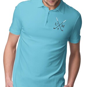 K&ouml;nnte beinhalten: Ein hellblaues Poloshirt mit wei&szlig;em Kragen und einem kleinen gestickten Design auf der linken Brust. Das Design zeigt zwei gekreuzte Golfschl&auml;ger mit den Buchstaben "U" und "M" dazwischen.
