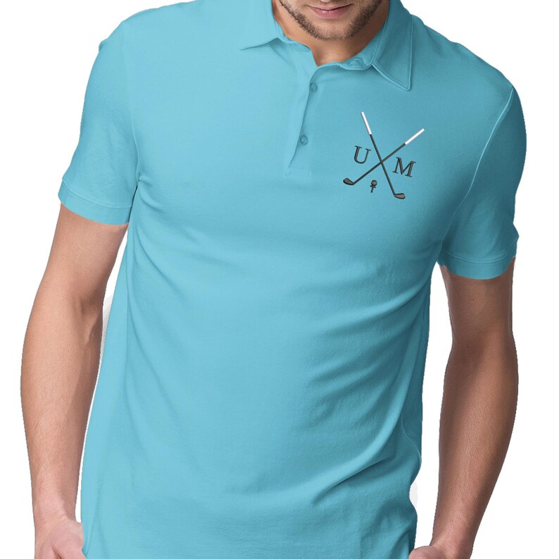 K&ouml;nnte beinhalten: Ein hellblaues Poloshirt mit wei&szlig;em Kragen und einem kleinen gestickten Design auf der linken Brust. Das Design zeigt zwei gekreuzte Golfschl&auml;ger mit den Buchstaben "U" und "M" dazwischen.