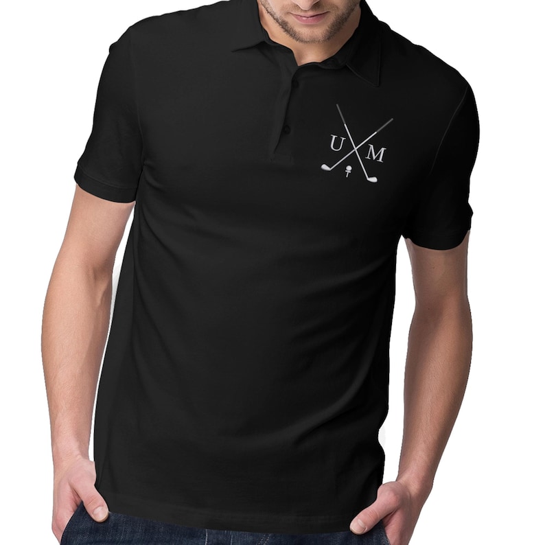 K&ouml;nnte beinhalten: Schwarzes Poloshirt mit einem wei&szlig;en gestickten Design aus zwei gekreuzten Golfschl&auml;gern und den Buchstaben "UM".