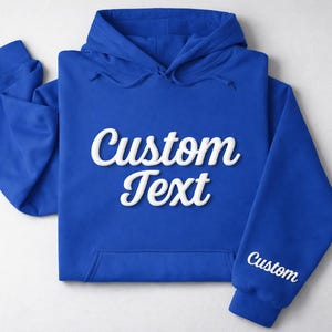 May include: Royal blue hoodie with the words "Custom Text" in white script across the front. The sleeve also has the word "Custom" in white script. The hoodie is laid flat on a white surface.